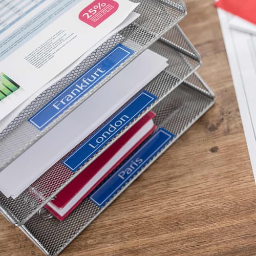 Bandejas metálicas de escritorio con carpetas, etiquetadas con los destinos Frankfurt, Londres y Paris usando la cinta adhesiva TZe-545 color azul letras blancas de la marca Brother para rotuladoras P-Touch