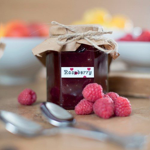 Frasco de mermelada de frambuesas etiquetado con cinta adhesiva blanca y fondo de corazones rosados TZe-MPPH31 de BRother.